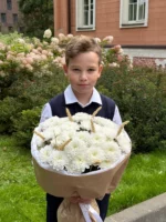 Является изображением мальчика, держащего букет из белых цветов на улице возле жилого дома.