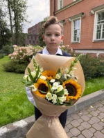 Ребенок с букетом подсолнухов и белых цветов на улице у кирпичного дома.