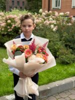 Ребенок с букетом розовых и персиковых цветов на улице перед цветущими кустами.