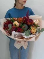 Большой букет из разноцветных цветов и зелени, фокус на женщине в синей куртке.