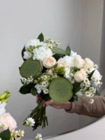 Букет из белых и кремовых цветов с лотосами и ромашками на фоне белой стены.