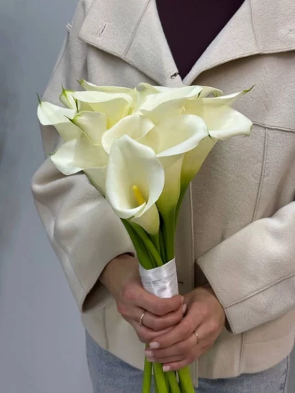 Букет белых калл в руках женщины на фоне серой стены.