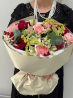 розовый и красный букет цветов с зеленью и пистелли, женщина в чёрном платье и жемчужным ожерельем.